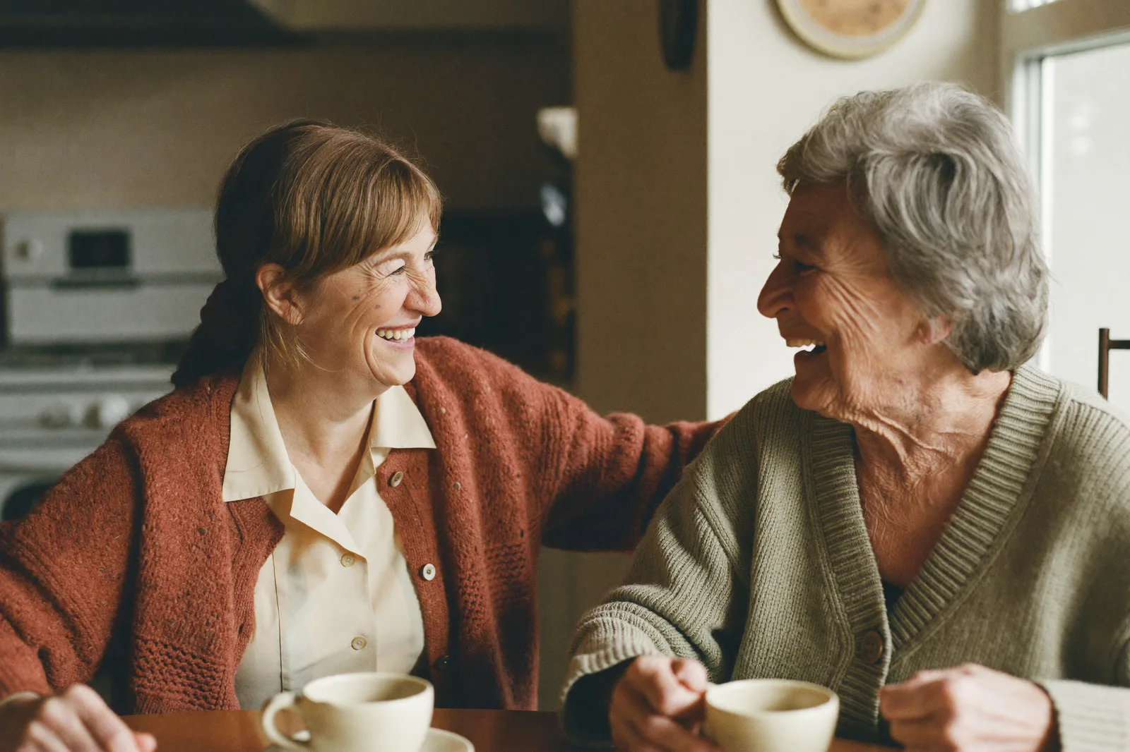 Alltagshelferin und Klientin lachen gemeinsam beim Kaffee am Küchentisch